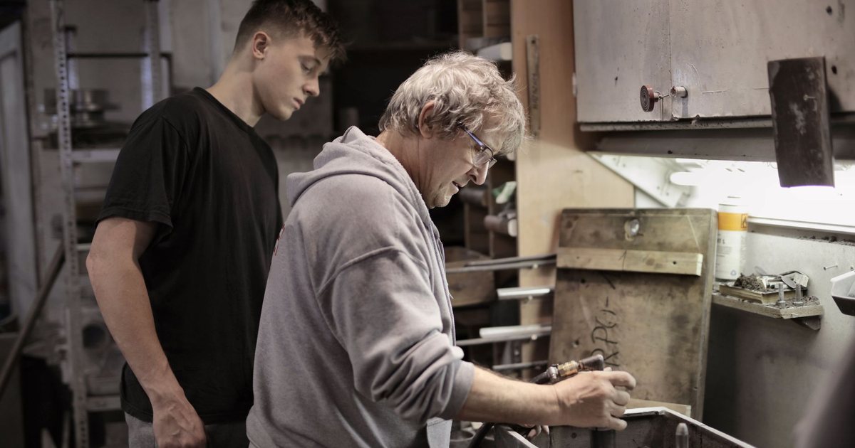 An older craftsman showing a young apprentice how to work a piece at a workshop bench