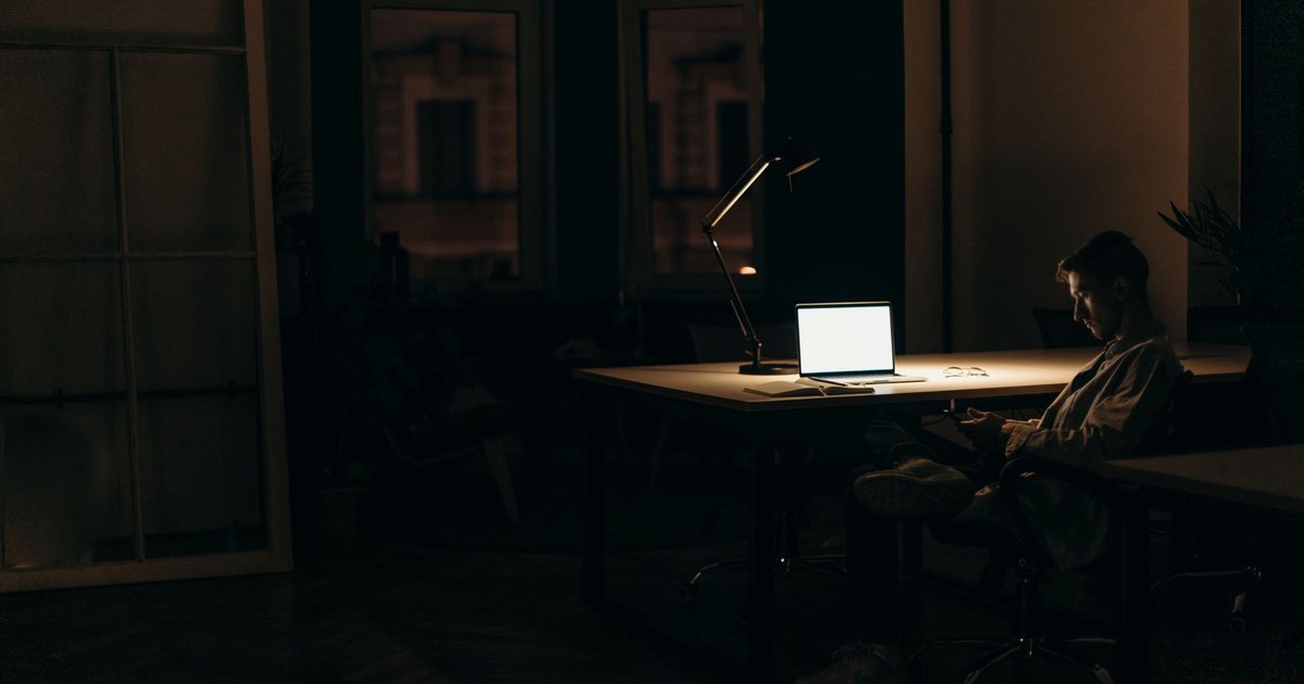 A person working alone at a laptop in a dark room, lit only by the screen and a desk lamp